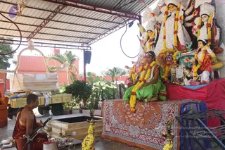A photograph of a Durga puja set up under a corrugated metal shelter with a red wall and palm trees behind. Beside a square stone yajna pit, a shaved-headed priest in orange-red dhoti and shawl sits cross-legged before a microphone, facing the shrine. A young girl in a green sari with gold border, decorated with marigold garlands and a gold headpiece, sits on a red and white embroidered cloth in front of a row of clay deity murtis, including a multi-armed goddess in white with a lion. Brass oil lamps with flowers and a tray of offerings sit on the ground. The scene looks like a puja or Navaratri worship rite.