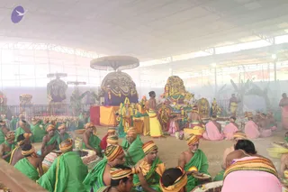 A large open-sided pavilion filled with hazy smoke or incense, where many men sit on the ground in green dhotis and green shawls with striped orange-and-yellow turbans. Some wear pink or magenta cloths beside them. Several people sit around a central altar with decorated deity murtis, a tall ornate peetha/throne draped in purple and gold, and a canopy with fringed gold trim. Bronze lamps and offering trays, a large cake, and pots sit on the floor. A bare-chested man in a yellow dhoti stands near the altar and another stands at right. The setting appears to be a ritual gathering, possibly a puja or abhishekam. No individual can be confidently identified as a person from appearance.