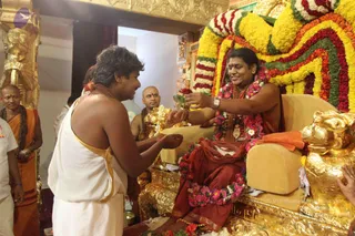 A seated man on an ornate gold throne with a carved gold lion armrest receives a red rose from a devotee in a white dhoti with gold border. The seated man wears a red-maroon shawl, a rudraksha mala, a gold pendant and a long garland of pink and orange flowers, and smiles. Behind the throne a large floral backdrop of yellow, orange, red and white marigolds and roses frames the seat. Other devotees stand nearby, including a shaved-headed priest in saffron and a man in white. Flower petals lie on the floor. a person, identity uncertain from appearance.