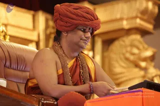 A man seated in a brown leather armchair wears a large orange-red turban, a saffron-orange shawl with gold trim, and multiple strands of beaded rudraksha-style malas with a gold ceremonial necklace. He leans forward at a dark lectern with an orange cloth, reading or writing, with a small orange cushion in front and ornate gold carved panels behind him. Swamiji (probable) is shown in appearance-based cues: orange turban, saffron clothing and rudraksha malas, while seated as a speaker at a stage lectern.