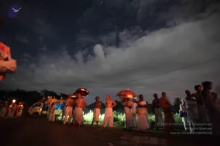 A night-time outdoor photo on a dark, muddy road beside a green field under a cloudy sky with a faint star. A line of about a dozen people in white dhotis and white garments stands in a loose row, several bare-chested or wearing sacred threads, while some hold red and orange umbrellas. A small white van and blue tarpaulins sit at left, with a few figures and lights behind. Blurred figures at right suggest a procession pausing on the roadside. No individual can be confidently identified, so no a person candidate is claimed.