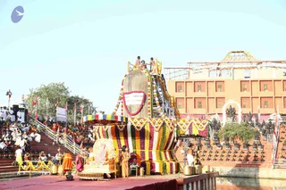 Outdoor ceremony at a temple-like courtyard with tiered stone steps lined with seated devotees in white and orange. A large stone lingam-shaped structure draped in multicolour flower garlands and stripes of red, yellow, green and blue stands at centre, with several young men in orange and bare-chested attendants on its top platform. In front, a golden throne with a radiant backrest holds a seated figure in orange robes, flanked by attendants and brass pots and lamps on a red stage. Black deity murti statues line the tiers on the right, and an orange building with a sloped roof sits behind. Clear blue sky overhead.