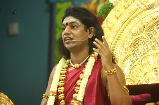 Photograph of a man speaking into a headset microphone during a satsang, gesturing with his right hand raised. He has long dark hair, a red bindi and white vertical lines on his forehead, and wears a red-maroon shawl draped over a bare upper body. He is adorned with a long garland of white jasmine and orange flowers, a double strand of large gold beaded necklaces, a silver chain, and gold bangles. Behind him is an ornate gold-embossed throne with floral green and red decoration. Probable Swamiji (probable) is seated on the throne, identified by long swept-back hair, rudraksha-style gold beaded malas and ceremonial gold ornamentation.
