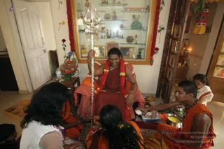 A photograph inside a home room decorated with red and orange flower garlands on the wall frame and a glass display cabinet holding small deity figurines and brass items. A seated man in a red-maroon robe and pink-orange shawl, wearing a red and yellow flower garland and gold bangles, holds a tall silver trident in his right hand while smiling and seated on a cushion. Around him, three people in orange and saffron clothing sit on the floor performing a pada puja, one pouring water or milk from a small cup, another with long dark hair attending, with a brass bowl of yellow flowers and a plate of offerings nearby. A young child in white stands at right, and a small vase of red and orange flowers sits on a side table. Robed devotees attend this house-visit ritual.