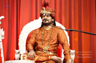 A man seated on an ornate white throne with carved lion-armrests in front of a deep red draped backdrop, speaking into a microphone on a boom stand. He wears a saffron-orange turban topped with a red plume and a gold jewelled headpiece, saffron-orange silk kurta, a gold necklace with a rudraksha mala and a gold deity pendant, and a gold waist belt, with a gold hoop earring. A small framed portrait sits on the throne's base at lower left.