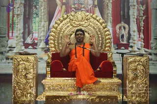A photograph of a seated man on an ornate gold throne with a red velvet cushion, dressed in orange saffron robes and wearing long rudraksha malas around his neck, raising both hands in a blessing gesture before a microphone. A large backdrop shows a decorated temple scene with a deity image in red and white, brass lamps and a small altar. Flower petals lie at the base of the dais. A wrap-style orange robe and bare feet are visible. a person is the central figure in an appearance-based label, consistent with the page's stated satsang talk.