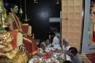 A photograph of a padapuja-style ritual in a temple room with black stone walls and a closed wooden door. A bare-chested man with swept-back dark hair, a white tilak on his forehead, a long orange-and-maroon wrap and a long garland of beads sits on a gilded throne with a golden lion-form base, smiling and resting his bare feet on a tray of red flowers. Several devotees kneel around silver trays holding small silver bowls, red flowers and offerings: a woman in a green sari, a woman in white, an older woman with grey hair, and a man in a grey shirt touching the flowers. Brass oil lamps and hanging bells stand near the throne, and a colourful deity statue stands at the right edge. Two people are visible at the back among the offerings.