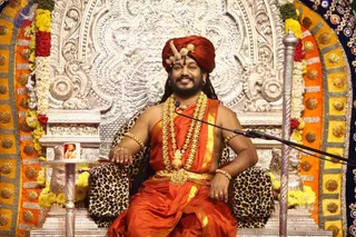 A smiling man with a long dark beard and long hair sits on an ornate silver throne decorated with a repoussé floral backrest, leopard-print cushions and yellow and orange cushioned trim studded with gold-toned knobs. He wears a red-maroon turban with a gold ornament, an orange-red saffron garment with gold border, a gold multi-strand necklace with a gold pendant, gold armlets and bangles, and a gold waist belt. A microphone on a stand is in front of him, and a framed portrait sits on a small silver side table beside him. Marigold and red flower garlands drape the throne. Presenting a Swamiji (probable) figure in a satsang setting, with the wearer seated on a throne in saffron-orange ceremonial clothing and a turban; the figure has long swept-back hair, a multi-strand gold necklace and gold armlets.