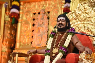 A seated man on an ornate gold-and-red throne smiles broadly at a microphone on a stand. He wears an orange-brown robe, a thick rudraksha mala, a ceremonial gold necklace with a large pendant, and a white-and-orange garland of jasmine and purple orchids. He has a white tilak with a red dot on his forehead, long dark hair and a bushy beard, and rudraksha bracelets on his wrists. Behind him are gold carved panels, orange decorations and hanging tassels in purple, red and white. Swamiji (probable): long swept-back hair, a bearded face, rudraksha mala and ceremonial gold necklace, seated on a throne as presiding teacher during a discourse.
