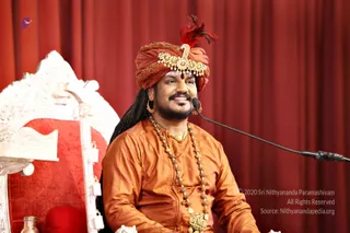 A smiling man with a trimmed beard and long dark hair sits before a red curtain, speaking into a microphone on a stand. He wears a red-orange turban with a gold ornamental brooch and a red feather, an orange-gold silk kurta, long rudraksha mala beads, a gold necklace with a pendant and gold earrings. Beside him stands an ornately carved white and red throne with a red velvet seat, and a metal vessel sits at the lower left. Appearance-based label: Swamiji (probable), seated in the speaker's position with distinctive turban, saffron-orange silk and rudraksha cues.