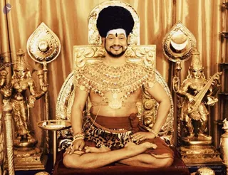 A seated man in a golden ornate throne sits cross-legged on a brown cushion before gold-toned draperies. He wears a tall black hair crown, white forehead markings with a red bindi, a layered gold necklace and chest ornaments, a gold waist belt, and a leopard-print lower cloth over an orange-brown wrap. Gold bangles are on his wrists. Flanking him are golden deity statues: a multi-armed figure on the left and a figure holding a veena on the right, plus a golden kalasha-style finial atop each side pillar and a small brass vessel near the throne. Swamiji (probable) is shown in the appearance-based label: a bare-chested man with a tall black crown, ornate gold jewellery and leopard cloth seated on a throne, matching the page's Swamiji reference. Photo credit text at lower right reads 2021 Sri Nithyananda Paramshivam