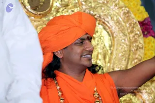 A man in an orange turban and orange shawl stands in front of an ornate gold-toned backdrop with yellow flower garlands at upper right. He has long dark hair falling past his shoulders, a red tilak on his forehead, and wears gold ornamental necklaces over the orange cloth. He smiles and raises one arm toward the right edge of the frame. A partially visible person in white clothing stands at the left edge. Swamiji (probable): the long swept-back hair, orange turban and shawl, and gold necklaces match the appearance of the Nithyananda Paramashivam candidate, shown in person.