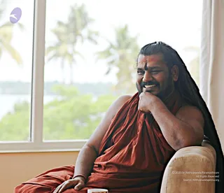 A man with long dark hair swept back sits in a cream upholstered armchair beside a large window, smiling broadly with his chin resting on his hand. He wears a rust-orange shawl wrapped over his shoulders and bare arms, with a dark vertical mark on his forehead. Through the window are blurred palm trees and water. A white cup sits on a surface in the lower foreground. Visible cues include long swept-back hair, orange clothing and a forehead mark; appearance-based identification as a person is plausible but not certain, so the candidate is marked unsure.