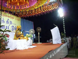 Night outdoor discourse on a raised stage with a red carpet. A seated figure in saffron-orange robes and an orange head wrap, with a gold necklace, sits on a white-draped chair behind a microphone and gestures with raised hands while speaking. Probable Swamiji by appearance: orange turban-style head wrap, saffron robe and ornate gold necklace, seated centrally as the speaker. Overhead, a canopy is decorated with orange and pink garlands and tassels. Bright stage lights hang at right, a white-draped lectern stands beside him, and two electric fans are placed on the stage. Several uniformed and white-clad attendants stand at the edge of the stage. Tamil banner letters are visible on a yellow and blue backdrop at left.