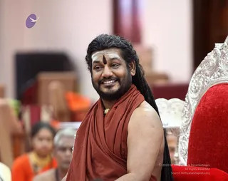 A smiling man with long dark hair swept back and a long beard sits in a hall beside a red velvet throne with silver carved armrest. He wears a sleeveless rust-red wrap over bare shoulders, a gold pendant at his neck and white sacred ash marks on his forehead with a dark central tilak. Blurred figures in orange and yellow garments sit behind him in the ritual hall during a Navaratri darbar. A small purple and white bird logo appears in the upper left.