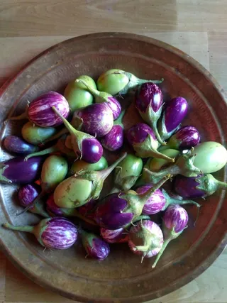 Photograph from above of a round, hammered copper or brass plate holding a heap of small brinjals (eggplants). Most are purple with white streaks, and several are pale green, with green calyxes and stems. They rest on a light wooden surface. No people or ritual setting are visible.