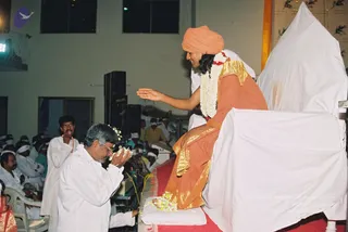 A seated figure on a white-draped throne with red carpet wears an saffron-orange turban, orange robe with gold shoulder trim, and a white flower garland with red accents, extending one hand toward a kneeling man in white shirt and trousers who holds flowers and bows his head. Behind them, seated devotees in white headbands and white clothing fill a hall with black speakers and a yellow-clothed attendant standing nearby. The seated figure shows distinctive saffron turban, long hair, orange robe and garland cues, so this is a probable Swamiji appearance.