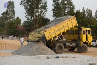 A yellow tipper truck, labelled with Karnataka state permit lettering, is tipping a load of grey gravel and sand onto a dirt and stone construction site. A man in a white shirt and light trousers stands at the left edge of the pile, and a few other figures are visible in the distant background near piles of stone. Tall eucalyptus trees and a low red boundary wall frame the scene, with a yellow plastic chair at the far left. No ritual, deity, or discourse is shown.