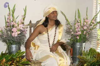 A seated man in white and gold ceremonial dress sits on an ornate carved wooden chair in a bright room. He wears a white turban with a gold border and a red bindi on his forehead, and a long rudraksha mala with dark brown beads hangs to his waist. His long dark hair falls past his shoulders, and he wears a white dhoti and shawl with gold borders. Tall pink gladiolus and lavender-toned flower arrangements stand in galvanized metal buckets on either side, with green croton plants in the foreground and window blinds at right.