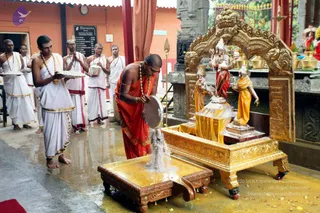 A ritual abhishekam at a temple shrine. A man in an orange-saffron dhoti and shawl, with rudraksha malas and a gold bracelet, pours water from a metal vessel over a deity murti of a seated figure on a gold-painted throne-shaped stand, with a ceremonial lid-plate beside him. Behind him, several men in white dhotis and bare upper bodies, some with sacred threads and shawls, stand holding silver plates and bowls of offerings. A golden arched torana frames the murti, with brass kalasha pots, flower petals on the ground and a red-and-gold canopy behind. Wet floor and a bulletin board reading the temple name are visible in the background.