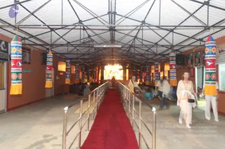 Interior photograph of a temple hall with a corrugated metal roof on steel trusses and orange-painted walls. A red carpet runs down the centre between metal railings toward a brightly lit shrine with a deity murti at the far end, with seated devotees on the floor in front. Hanging cloth banners in red, blue, green and yellow with floral and bird motifs line both sides. Two visitors in white clothing walk along the carpet, a woman in a light sari-style wrap with a handbag and another woman in white trousers, while others sit near the shrine. Framed portraits hang on the right wall.