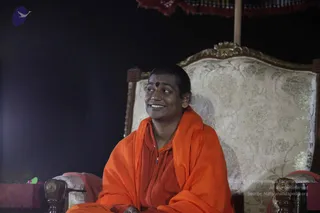 A smiling man with a short cropped hairstyle and a red bindi sits on an ornate cream upholstered throne with gold floral patterns and a gilded frame, under a red fringed canopy. He wears a saffron-orange shawl draped over an orange zipped top and looks off to the side against a dark background. A small purple bird logo appears in the top left corner. The image is a photograph of one person.