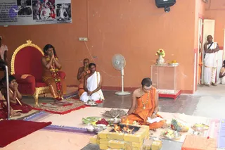 A homa fire ritual in an orange-walled hall. A priest in saffron robes and a long rudraksha-style necklace sits on the floor before a brick fire pit with burning wood, reading from a paper while offerings of fruit, flowers and brass bowls surround him. Behind, a seated man in white dhoti with a shawl holds a ritual vessel, and another saffron-clad man sits with hands folded. On a red and gold throne to the left sits a person in orange garments and a flower garland with hands joined in prayer, with two children in red-and-gold sitting nearby. A printed photo collage with a caption hangs on the wall above. A fan and a glass-cased pedestal are visible at right.