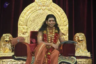 A photograph of a seated man on an ornate golden throne with carved lion heads on each armrest, set against deep red velvet drapes on a stage. He wears an orange-saffron dhoti and shawl, long flowing dark hair, a red dot on his forehead, gold arm bands, and a thick multi-strand necklace of red and green beads with gold ornaments, and a rudraksha mala. He sits with a microphone at his ear, smiling, one arm resting on the armrest. Presentation cues consistent with the page are present, so this is an appearance-based a person seated in a teaching setting, with a small brown ceremonial cushion beside him.