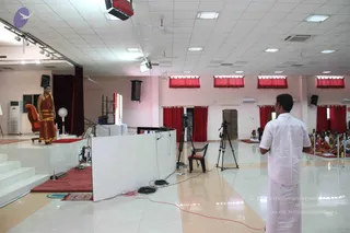 A large bright hall with white tiled floor and pink walls, red curtains on windows, and a raised white stage at left. A woman in a red and gold sari stands on the stage beside an empty orange-brown chair and a red cushion. In the foreground a man in a white shirt and white dhoti stands with his back to the camera, facing the stage. A video camera on a tripod and another tripod with a monitor are set up near a small white partition. Seated devotees in white and orange clothes sit on red mats at the right side of the hall, with a few people visible in the background. Ceiling lights, air conditioners and a standing fan are visible.
