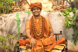 A man seated cross-legged on a wooden chair draped with an orange cloth and a tiger-striped mat, in front of a sculpted rock backdrop with golden seated figures in namaskar pose and green foliage. He wears an orange turban, orange shawl and dhoti, a brown rudraksha mala and a gold bead necklace, with gold bangles on his arms and a red tilak on his forehead, and a small headset microphone is visible. He smiles toward the camera with his hands resting on his lap. Appearance-based label, Swamiji (probable): the orange turban, saffron robes, rudraksha mala and gold necklace are the qualifying cues, and he is physically present as the seated speaker of a satsang setting.