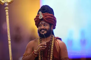 A smiling bearded man in a rust-red turban with a jeweled brooch and long dark hair piled above it stands on a stage, holding a small headset microphone near his mouth. He wears a rust-red shawl over bare shoulders, layered rudraksha malas and a gold-toned ceremonial necklace, gold armlets and a gold bangle. A tall gold staff with a decorated top stands at left against a warm yellow-and-blue blurred backdrop. a person: the turban, long hair, ornate gold necklace and rudraksha malas are visible cues of the speaker's role, a discourse on stage.