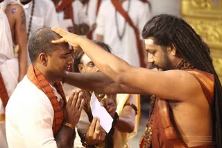 A photograph of a ritual blessing in a temple hall. A man with long dreadlocks, bare-chested, wearing a rust-orange and gold-bordered shawl and a rudraksha mala, presses his palm on the forehead of a shaven-headed devotee in white dhoti and an orange-red scarf printed with script. The devotee stands with palms joined in anjali. Behind them a priest in white and orange clothing holds a folded paper, and other white-clad participants with red scarves stand in the background beside gold-coloured walls. a person: the dreadlocked, bare-chested figure in orange-red shawl with rudraksha beads and a white forehead mark is applying a blessing, consistent with Pada Puja or initiation rites.