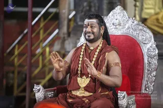 A photograph of a man seated on an ornate silver throne with a red velvet cushion and carved lion-head armrests, smiling with palms raised in a gesture of address. He wears a rust-red saffron-toned cloth wrapped over his shoulders and torso, a long ornate gold necklace of round beads, a gold pendant with a temple-tower motif, a gold waist belt, gold armlets and beaded bracelets, and a white forehead mark with a red dot. Long dark hair falls behind him. Behind the throne are gold-painted and metal-railed structures. Appearance-based probable Swamiji (probable) seated in the speaker role, with long hair, saffron-toned clothing and ceremonial gold necklaces as visible cues.