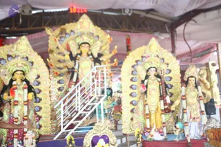 A photograph of several large decorated deity murti standing on a raised platform under a pink and white canopy in a temple-like hall. The central figure is a multi-armed goddess with a golden halo and crown, holding a weapon in one raised hand and a lotus-like object in another, flanked by two ornate arches. To the left and right stand other goddess murti with garlands of white and red flowers, one holding a veena-like instrument. A white metal staircase runs up the platform in front, and small brass lamps and flower offerings sit at the base. Ornate golden arches frame each figure. Ceiling lights and a red digital clock appear overhead.