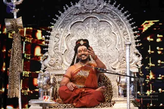 A seated man in a long-haired, crown-adorned look sits on an ornate silver throne with carved elephant-motif arms, gesturing with his raised right hand while speaking into a microphone on a stage. He wears a rust-orange wrap and a leopard-print shawl over his shoulder, with rudraksha beaded bracelets and a gold necklace. A silver ceremonial staff with a leopard-print cloth stands to the left, and a small framed portrait is placed at the stage's lower left. Background screens glow with red and orange lights. a person: the appearance cues of a swept-back long hairstyle, ornate silver throne, leopard-print and saffron-orange ceremonial clothing, rudraksha malas and gold necklace match the discourse setting; the figure is shown as a speaker giving a talk on stage.