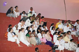 A photograph of a large seated audience on tiered red-brick steps at dusk, with a pink sky behind them. Most attendees wear white ritual clothing, with some in colourful saris, shawls and salwar kameez. Many sit with eyes closed or hands folded in prayer, and one man in an orange-and-white outfit with an orange shawl sits near the top. A video camera on a tripod is at the lower right. The scene matches a satsang or talk, but no speaker is visible. Roughly 40 people are visible.