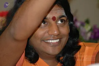 Close-up photograph of a smiling man with long, swept-back black hair and a red dot on his forehead, wearing an orange-saffron garment. One arm is raised across the upper frame, and blurred purple flowers appear in the background. The man appears to be a person, based on the long dark hair, saffron clothing and forehead mark; he is shown in a close, candid pose.