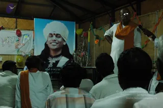 A standing man in a white shirt and white dhoti with an orange saffron shawl over his shoulder speaks into a handheld microphone to a seated audience in a thatched bamboo hall. Behind him a large framed portrait labelled with the printed caption "Paramahamsa Nithyananda" shows a smiling figure in a white turban with a red bindi, a dark robe and a layered pearl necklace. Colourful paper streamers, balloons and a banner with cut-out letters decorate the walls. Several seated men in white and orange shawls face the speaker, their backs to the camera.