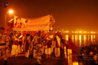 Night procession at the riverside, with orange-lit water and distant lamps behind. A large white banner carried by marchers shows a portrait of a figure with long dark hair and a red garment beside printed Nithyananda Dhyanapeetha text. Marchers in white dhotis and red sashes walk barefoot, while uniformed guards in camouflage flank the procession and carry rifles. Red flags and a decorated ceremonial pole rise above the group.