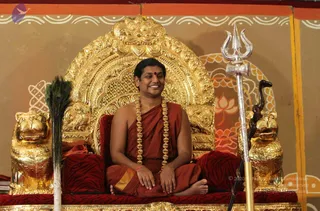 A smiling man in saffron-red robes sits cross-legged on an ornate golden throne with a carved arched back and gilded lion figures on each side. He wears a long ceremonial gold beaded necklace and a red bindi, and his hands rest on his lap. A silver trident stands to his right and a peacock-feather fan to his left, with a dark serpent-headed staff beside the trident. The red-and-black tiger-striped throne cushions and a painted orange and brown backdrop with lotus motifs complete the setting. Appearance-based label: a person, seated on a throne, based on the saffron robes, ceremonial gold necklace and throne setting.