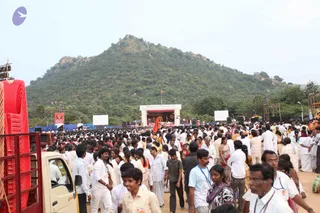 A large outdoor crowd fills a dirt ground in front of a stage at a Nithyananda Dhyanapeetam event, with a forested hill rising behind. Many people in white dhotis and shirts, women in saris, and volunteers wearing lanyards walk and stand through the crowd. A white arch on the stage reads Nithyananda Dhyanapeetam, and a saffron flag flies at the centre. A red truck and red chairs stand at the left edge, and a small red-and-white banner with a portrait is hung on a pole.