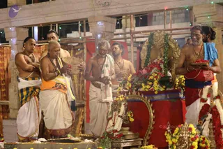Outdoor temple ritual at night beneath strings of multicoloured fairy lights along a pillared pavilion. Several men in traditional south Indian attire stand before an ornate golden deity murti or throne-like shrine heaped with red and yellow flower garlands. A bald man in white dhoti with a saffron-orange shawl and a rudraksha mala holds a woven basket of red flowers. Beside him a shirtless man with a grey beard and white dhoti with green-bordered cloth stands with folded hands. Another man in a red garment with blue cloth holds a tray of red petals, and white flower garlands hang at the right edge. Brass vessels and a lamp sit on the floor in front. The scene looks like an abhishekam or puja. None of the men is identifiable as a person from appearance cues shown here, so no candidate is asserted.
