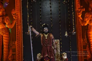 A man stands on a raised stage before a dark stone-panelled backdrop hung with strings of warm fairy lights and brass bells, flanked by large orange-lit sculptures of deities. He raises his right arm holding a tall silver trident, wears a maroon-and-gold dhoti-style cloth, layered rudraksha malas, a heavy gold necklace and a crescent ornament on his head, with white tilak marks on his forehead. A gilded throne with golden lion-head feet sits behind him. Oil lamps and a hanging bell frame the scene.