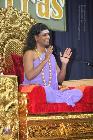 A man seated cross-legged on an ornate gold throne with red velvet cushions, speaking into a microphone with both palms raised in an expressive gesture. He has long, swept-back dark hair, a white and red forehead marking, and wears a lavender draped garment with a large gold bead necklace of rudraksha-style beads. The throne has heavily carved gold panels, and a blue curtain and green backdrop with gold letters are behind him. The appearance-based label is a person, seated in the teaching role of a satsang discourse.
