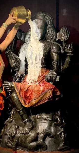 A dark stone murti of a seated deity with a trident-like staff and a crown-like headdress is being bathed with milk poured from a golden pot held overhead by a bare arm wearing bangles. The milk flows down over the figure's face and chest onto a red and gold draped cloth covering its lap. The murti rests on a carved pedestal showing small figures, and a dark background surrounds it. This appears to be a milk abhishekam of a deity murti.