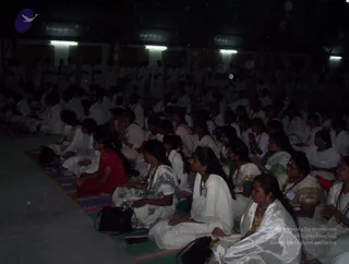 A dimly lit hall with many seated devotees, mostly women, sitting cross-legged on striped mats and facing toward the front. Many wear white or cream saris and rudraksha malas; one woman in the foreground wears a white sari with a gold border and a white shawl, another in a floral white sari, and one in a red top at left. Name badges are pinned to several clothes. Men in white stand at the back of the hall beneath fluorescent lights. The scene suggests a healers' initiation session with a meditative gathering.