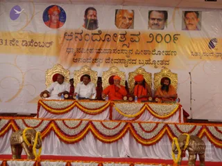 A stage event with five people seated on ornate gold thrones behind a flower-decorated table draped in white and orange-yellow marigold garlands. From left, two men in white shirts sit at the ends, followed by three figures in saffron-orange robes: the centre one in an orange cloth on his head, the second with long hair and a crown-like orange headpiece with a rudraksha mala, and the third with a long beard and orange robes. Gold ceremonial elephant figures flank the stage, and a backdrop shows portraits in circular frames above Kannada text. The printed name on the banner is a caption, not an identification of any seated person. The seated figure with long hair and orange headgear shows distinctive ceremonial cues, so the candidate is marked as probable, but the appearance is not conclusive enough to confirm identity.