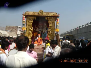 Outdoor photograph of a golden ratha (chariot) procession on a dusty road beside shops and a concrete flyover wall. The ornate gold-painted shrine on the chariot holds a deity murti of two seated figures, one with a trident and rudraksha mala. In front, a white-clad figure in saffron-orange robes with a red turban sits on a raised platform, hands raised in blessing, with a garland. Drummers in white and a man in a pink shirt stand near the platform. Dense crowds of men in white shirts and dhotis surround the chariot, with auto-rickshaws and trucks visible behind. Several figures are visible; crowd counts are approximate.