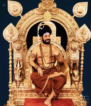 A bearded man seated cross-legged on an ornate gilded throne with lion figures and gold arch, bare-chested, wearing a tall dark turban with a crescent emblem, multiple rudraksha malas, gold necklaces and armlets, and rust-brown dhoti with gold border. He raises one hand in blessing. A trident stands at the left of the throne against a dark blue backdrop.