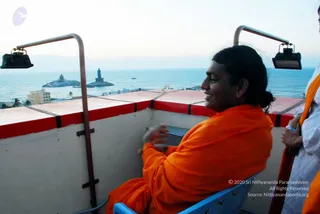 A photograph taken from a raised platform overlooking the sea. A man with long dark hair tied back, wearing an orange saffron shawl, sits in a blue plastic chair facing the water and smiles. A woman in white clothing with an orange wrap stands at the right edge. Behind them, the sea stretches to a horizon, with a rocky islet holding a stone monument and a tall statue on a second rocky outcrop, plus a low building and palm trees on the shore. Two metal lamps on curved poles frame the platform.